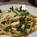 A beautifully plated dish of fettuccine pasta tossed in pesto sauce, topped with grilled chicken slices, fresh parsley, and grated Parmesan cheese, served on a white plate.